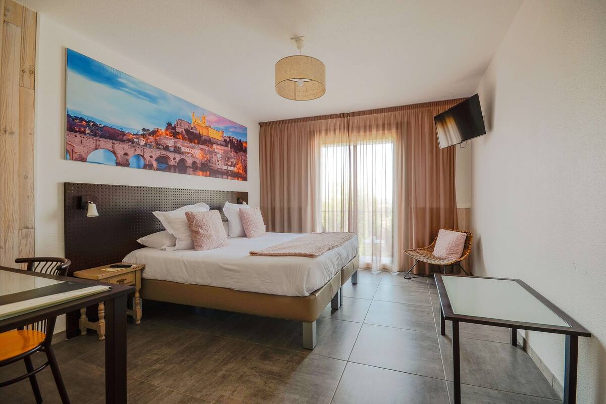 A hotel room with a bed, a desk, a chair, a TV, and a painting of a bridge.