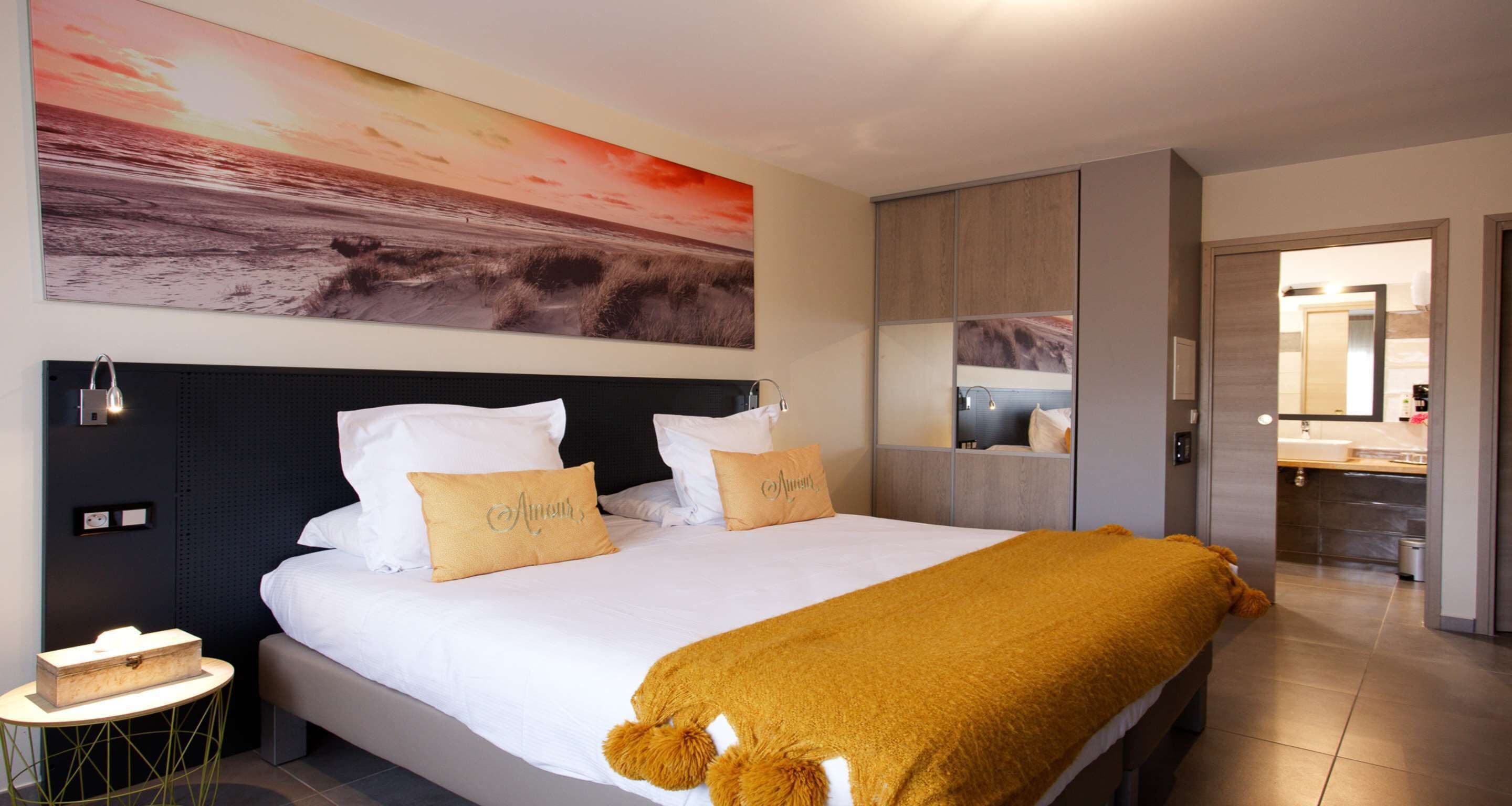 Une chambre d’hôtel avec un grand lit, une table de chevet, une lampe fixée au mur, un tableau représentant un coucher de soleil, un miroir et une salle de bain visible à travers une porte ouverte.