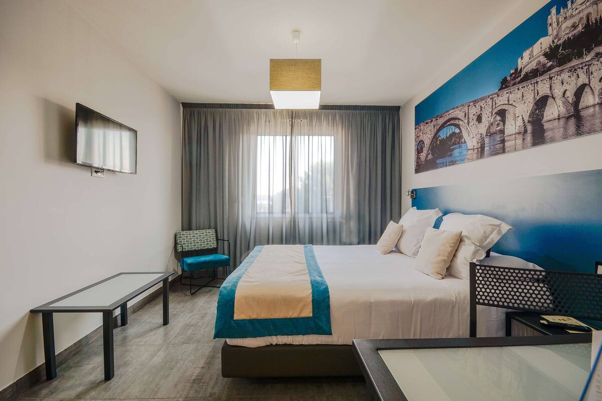 A hotel room with a bed, a desk, a chair, a TV, and a large window with curtains.
