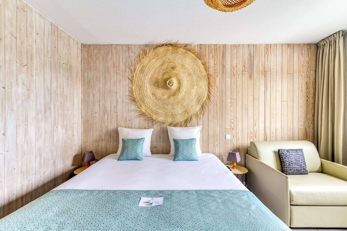 A hotel room with a bed, two pillows, a chair, a bedside table, and a woven wall hanging.