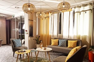 Habitación de hotel moderna con sofá, dos sillones, una mesa de centro y una mesa de comedor. Incluye lámparas decorativas, cortinas y una planta en maceta.