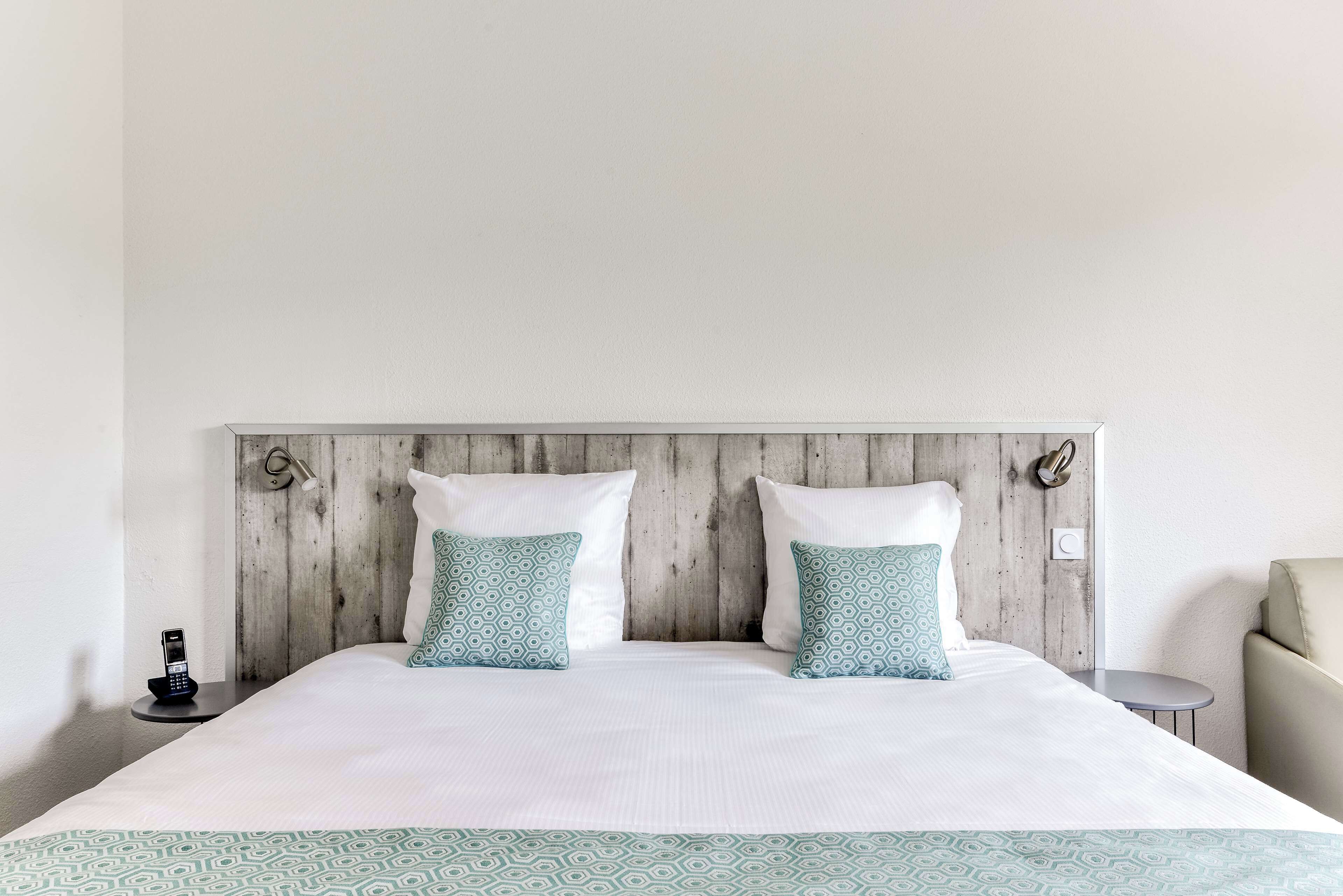 A neatly made bed with a patterned bedspread, two pillows with a geometric design, a wooden headboard, and a bedside table with a phone.