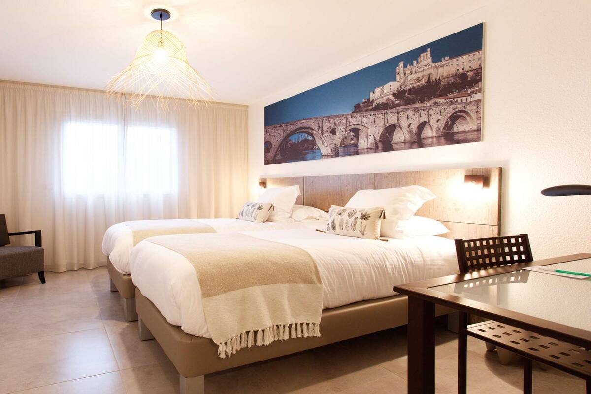 A hotel room with two beds, a desk, a chair, a TV, a large window with curtains, and a painting of a bridge.