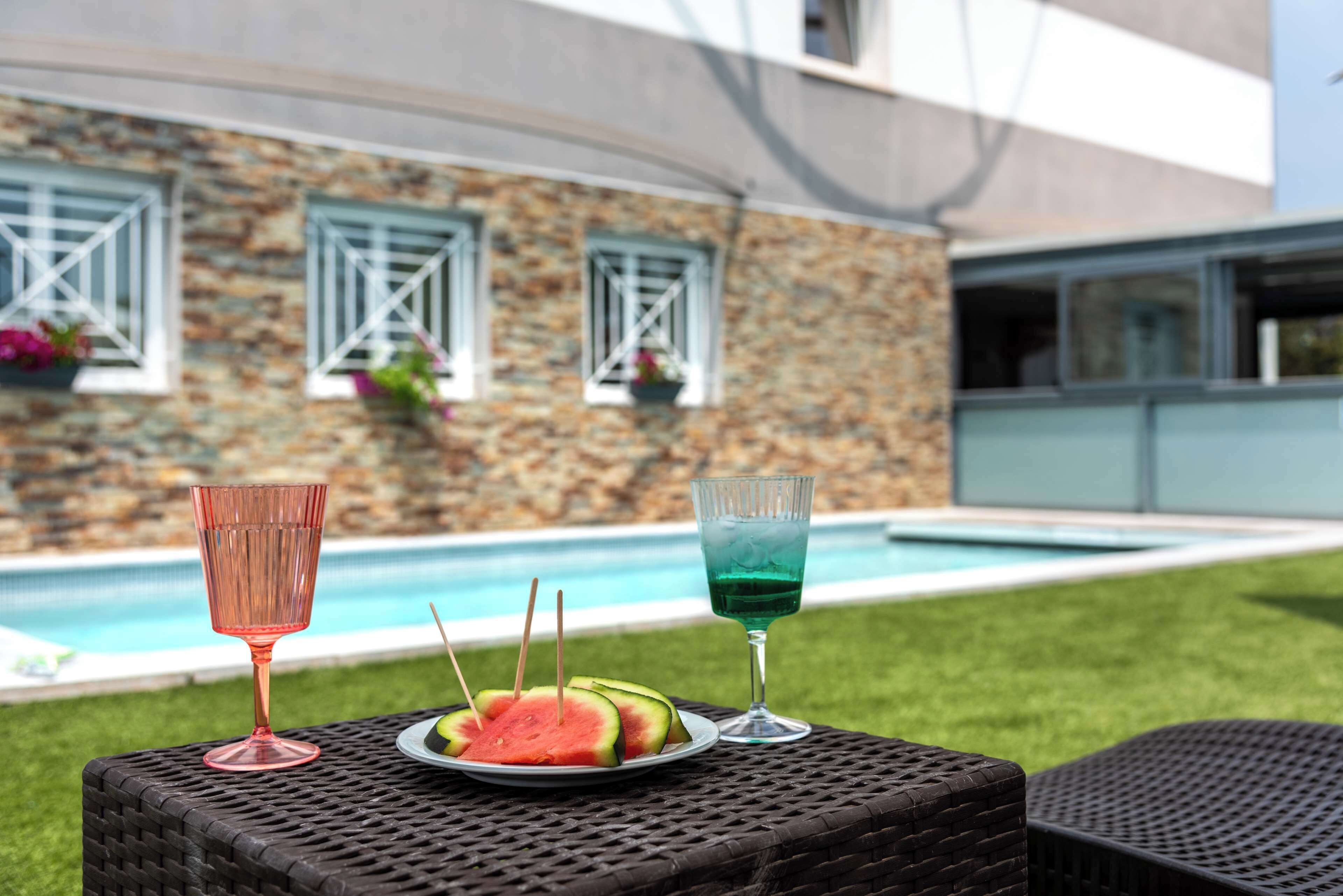 A poolside setting with a stone building, two drinks, and watermelon slices on a table.