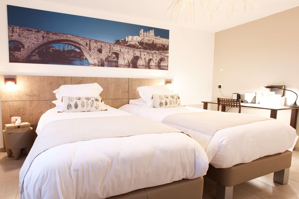 Une chambre d’hôtel avec deux lits, une tête de lit en bois, une table de chevet, un bureau et un grand tableau représentant un pont, accroché au mur.