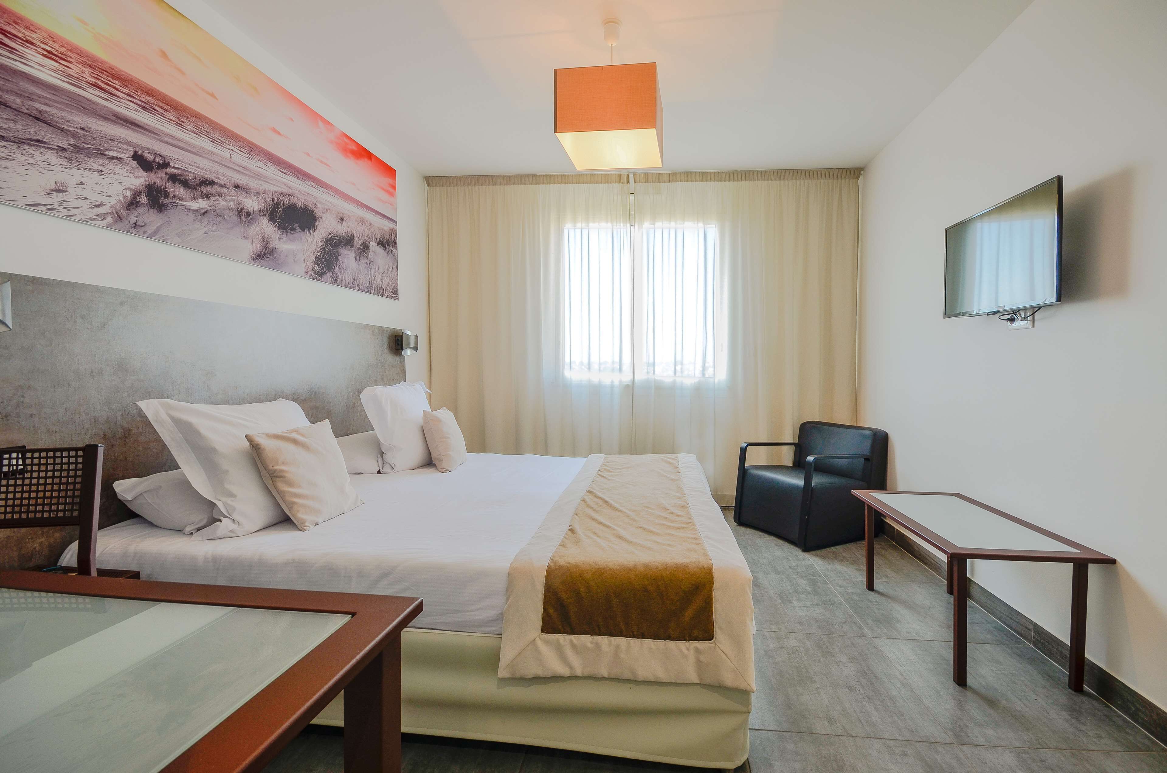 Quarto de hotel com cama, escrivaninha, cadeira, televisão e uma janela grande com cortinas.