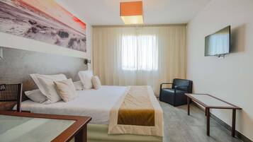 Quarto de hotel com cama, escrivaninha, cadeira, televisão e uma janela grande com cortinas.