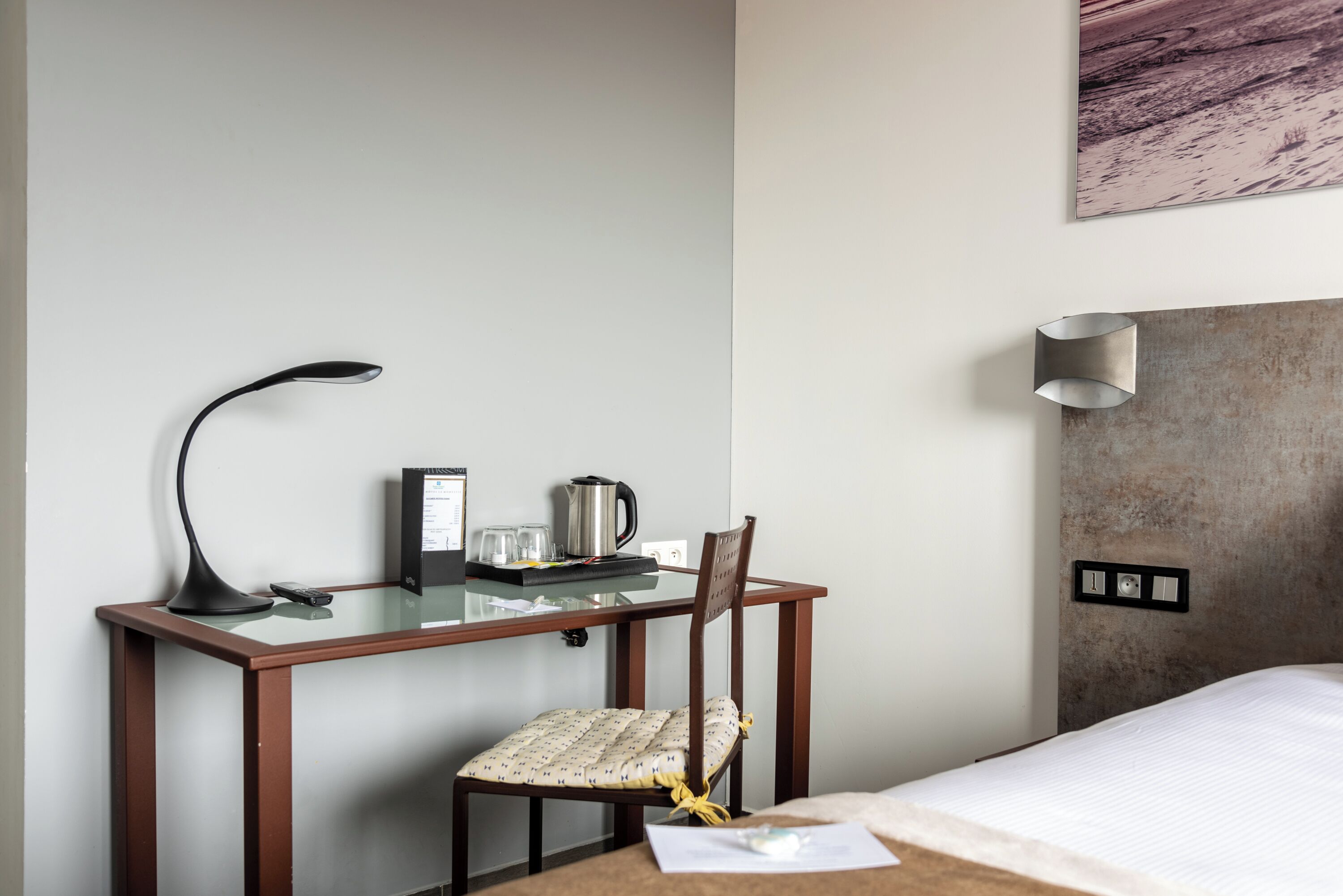 Una habitación de hotel con una cama, un escritorio con lámpara, una silla, una cafetera y una luz en la pared.