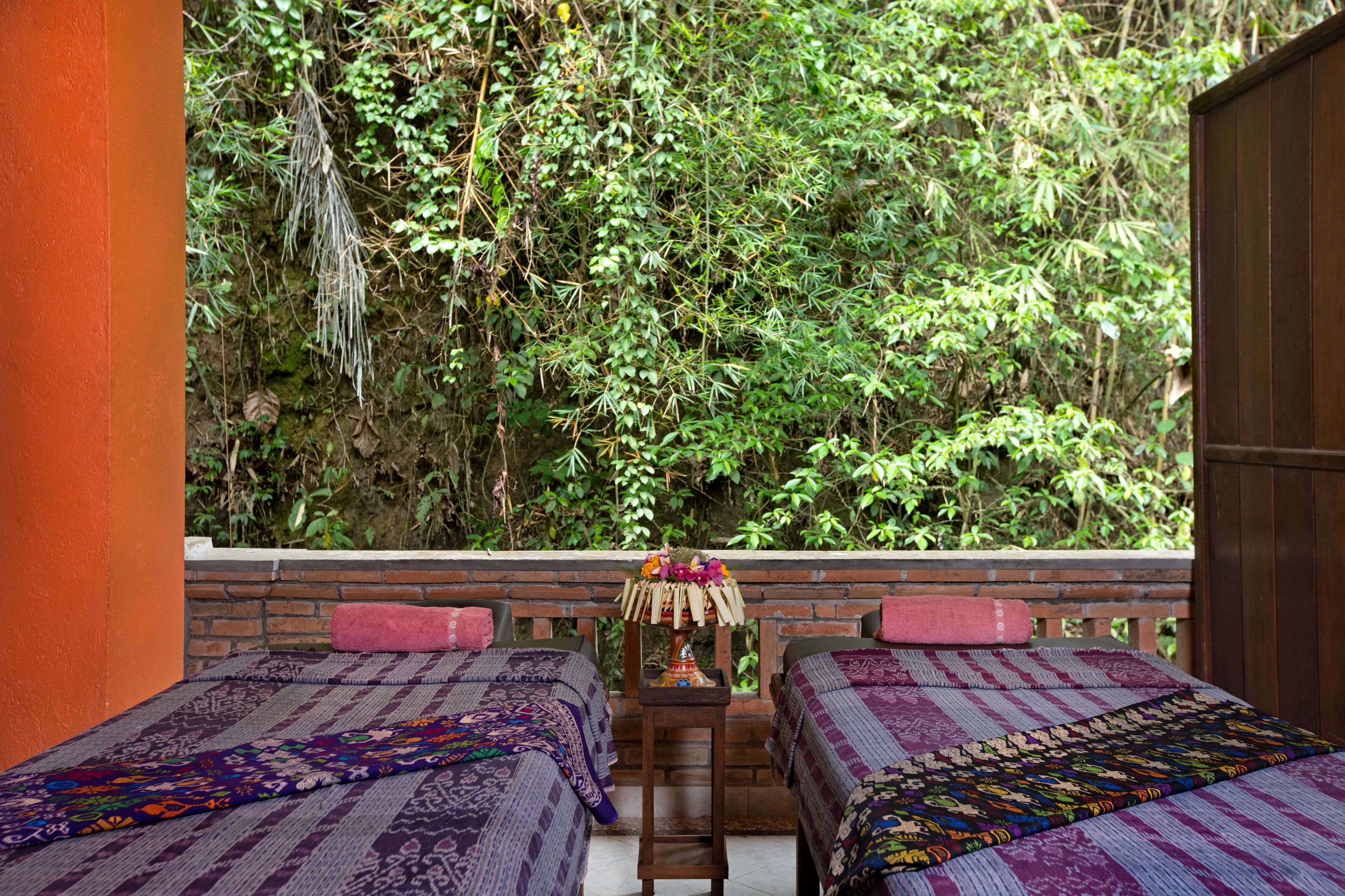 A room with two beds, a small table with a lamp, and a view of dense greenery.