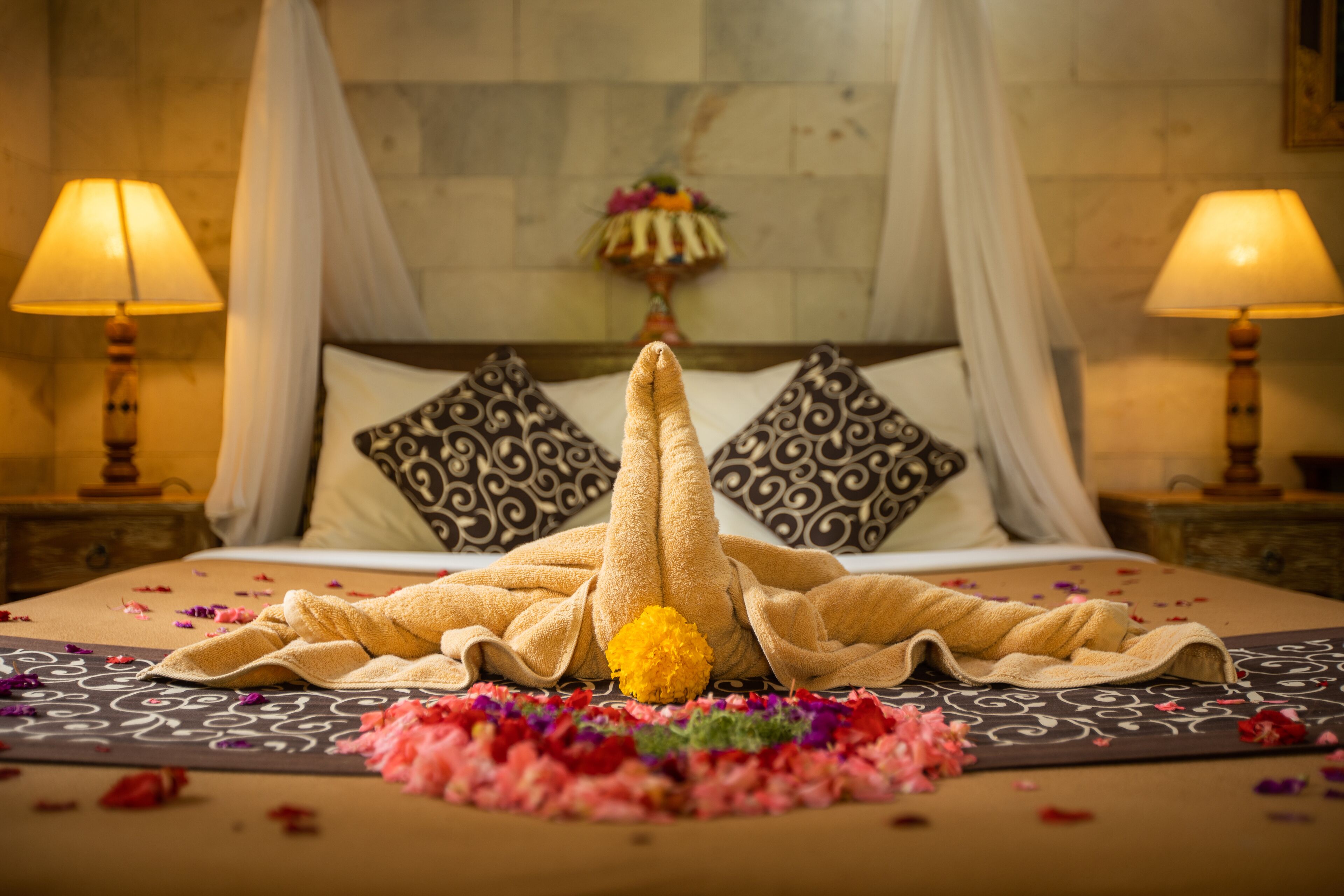 A neatly made bed with a towel and a yellow flower centerpiece, surrounded by scattered rose petals.