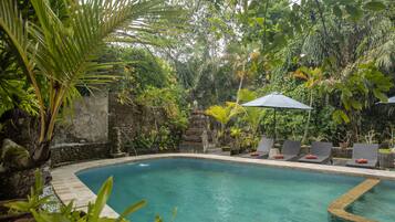 A pool with stone walls and tropical plants, surrounded by lounge chairs and a stone statue.
