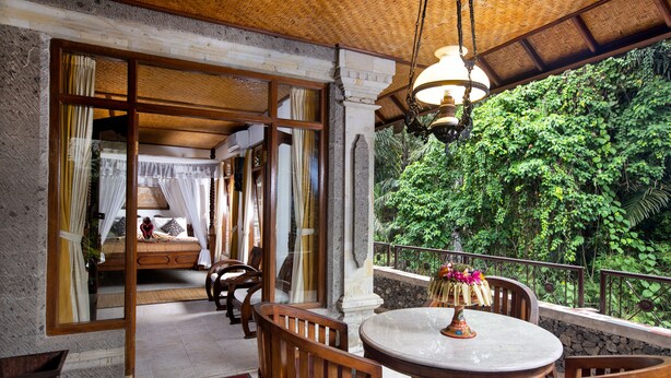 A room with a bed, wooden chairs, a round table, and a balcony with a view of lush greenery. - Ketut's Place Bed & Breakfast Ubud (Ubud)
