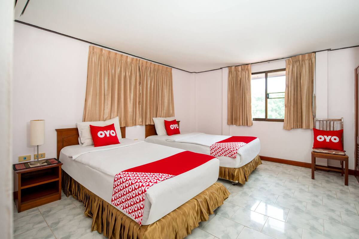 A hotel room with two beds, beige curtains, a window, and a nightstand with a lamp.