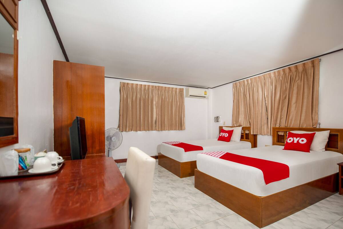 A hotel room with two beds, a wooden desk, a chair, a fan, and a small table with cups and a jar.