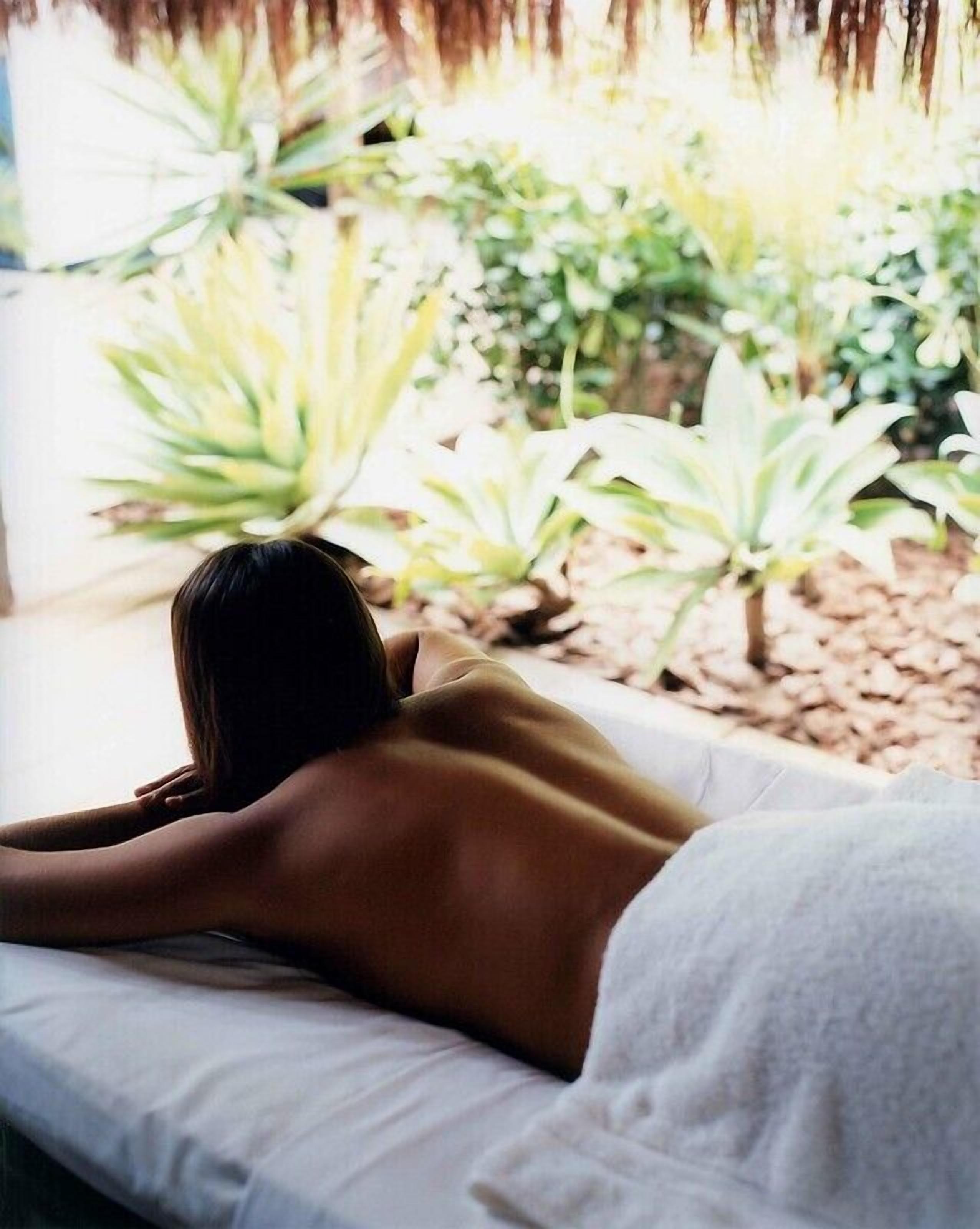 A person lying on a bed with a white sheet, in a room with a view of plants and a thatched roof.