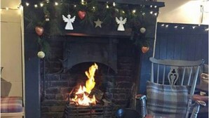 A fireplace with a fire, surrounded by Christmas decorations, a plaid chair, and a basket of wood. - The Queens (Carlisle)