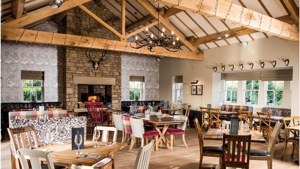A rustic dining area with wooden tables and chairs, a stone fireplace, and a high wooden ceiling with exposed beams. - The Queens (Carlisle)