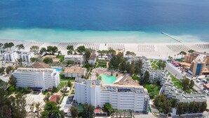 Vue aérienne d’un hôtel situé sur la côte, composé de plusieurs bâtiments, avec une piscine et une plage parsemée de parasols.