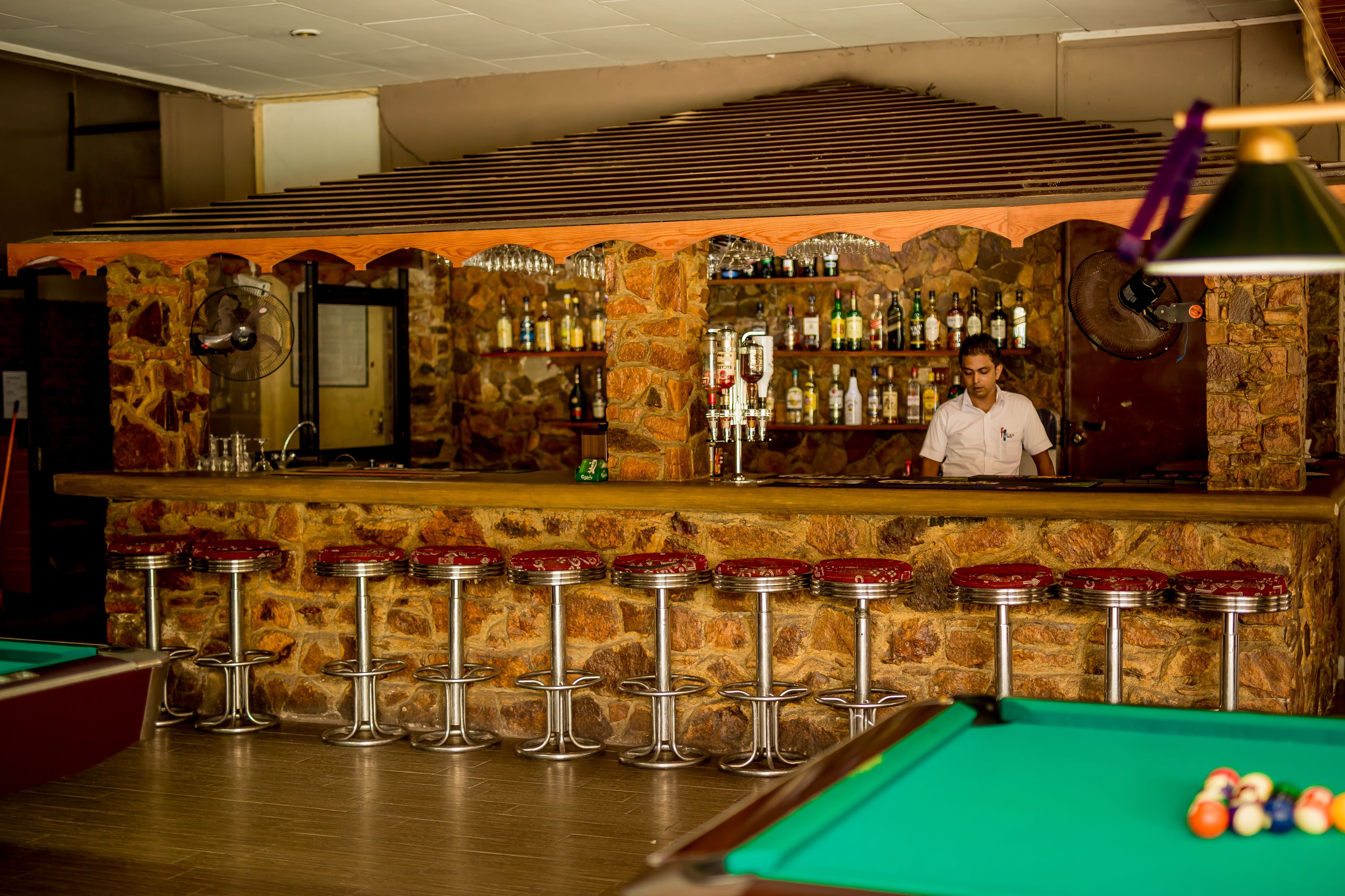 Zona de bar con un bartender, paredes de piedra y una mesa de billar.
