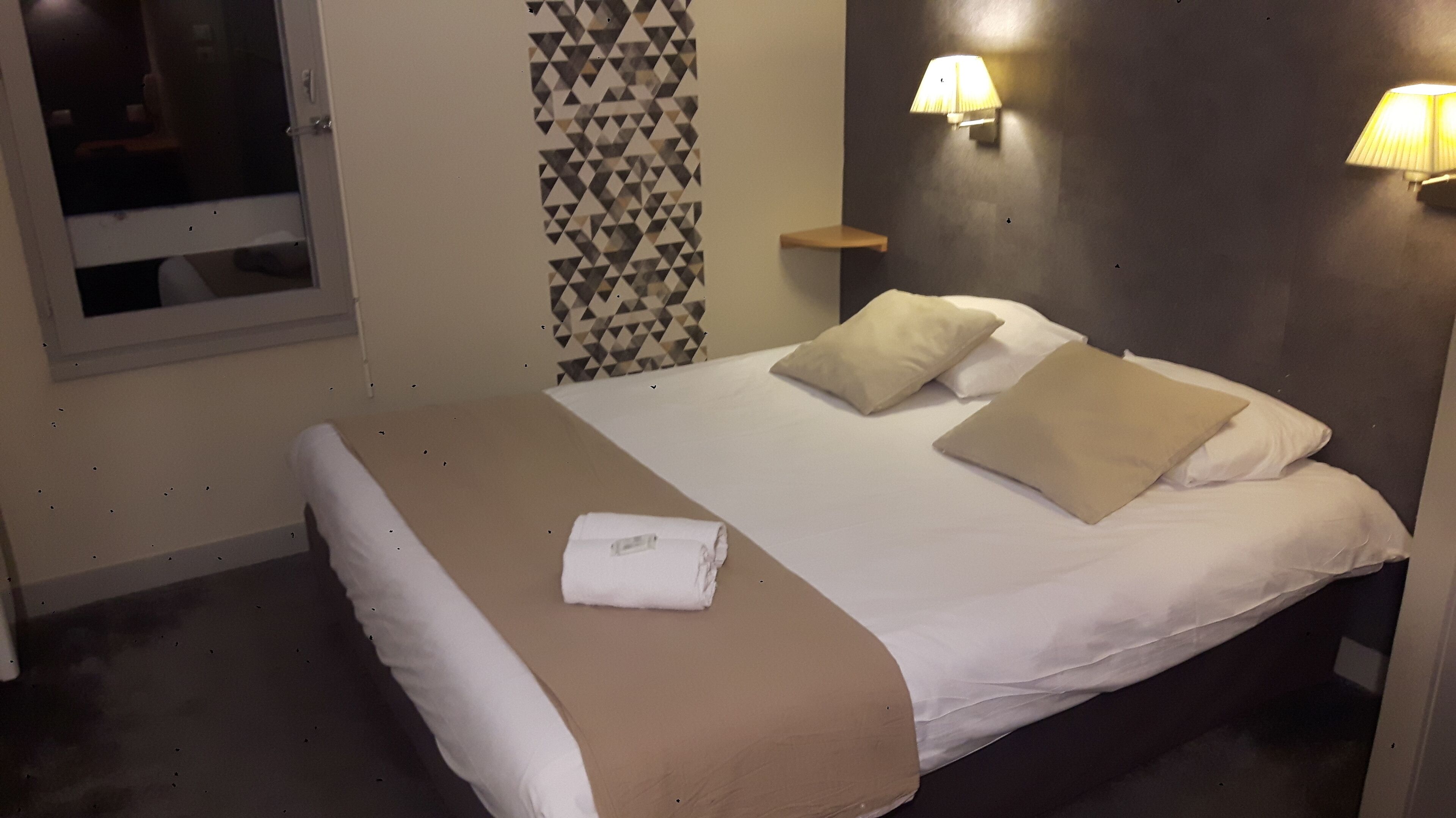 A neatly made bed with a brown blanket and white pillows, a small bedside table with a lamp, and a geometric-patterned wall.