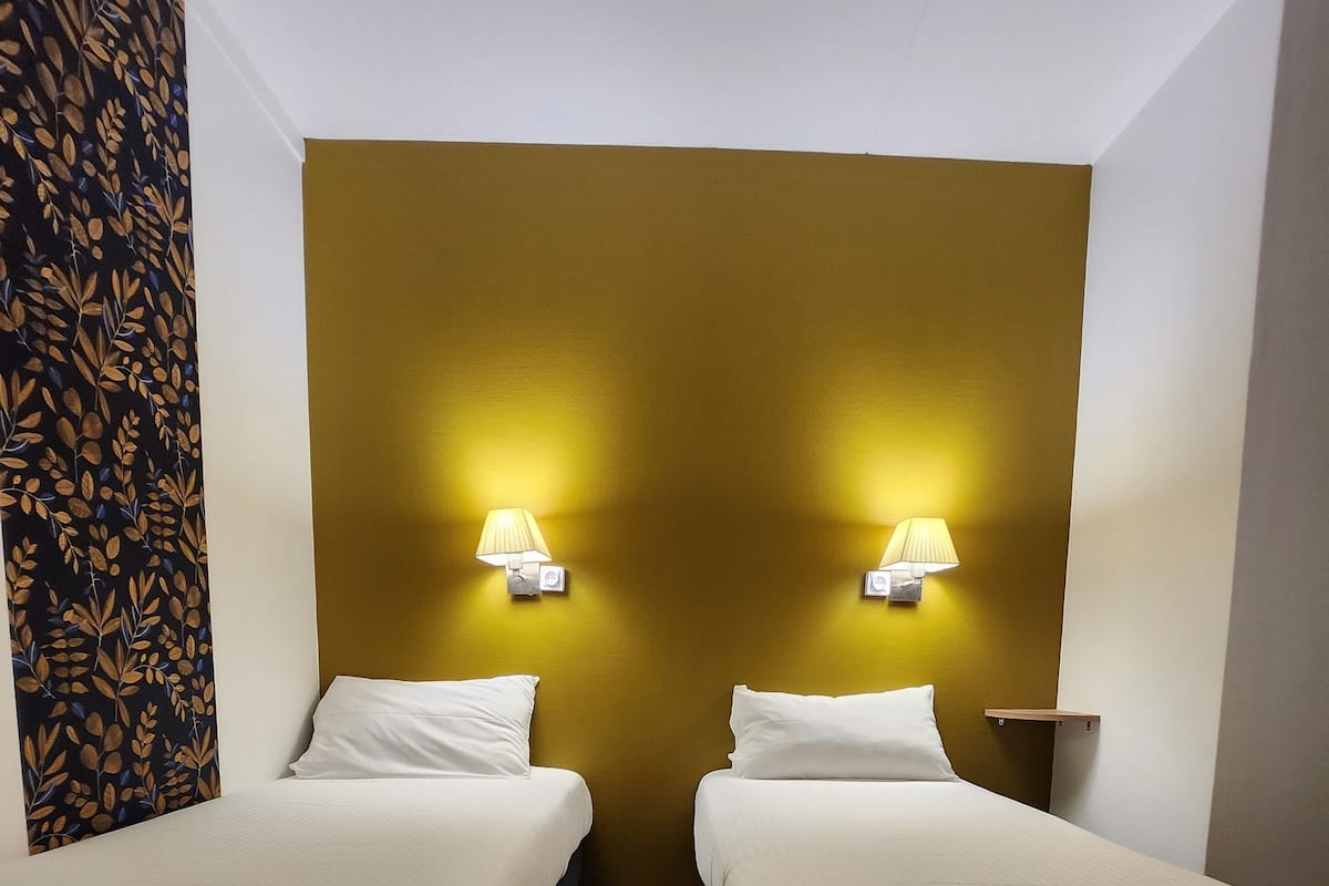 Ein Hotelzimmer mit zwei Einzelbetten, Holzboden, einer Wand in Senfgelb und zwei Wandleuchten.