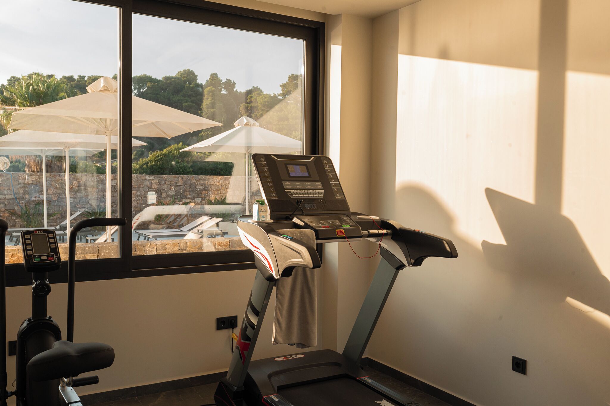 A gym with a treadmill, exercise bike, and a window with a view of outdoor umbrellas and greenery.