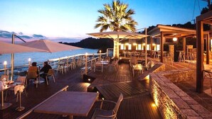 Une terrasse en bois, agrémentée de tables et de chaises, donnant sur une étendue d’eau, avec un palmier et un bâtiment abritant un bar visibles au loin.