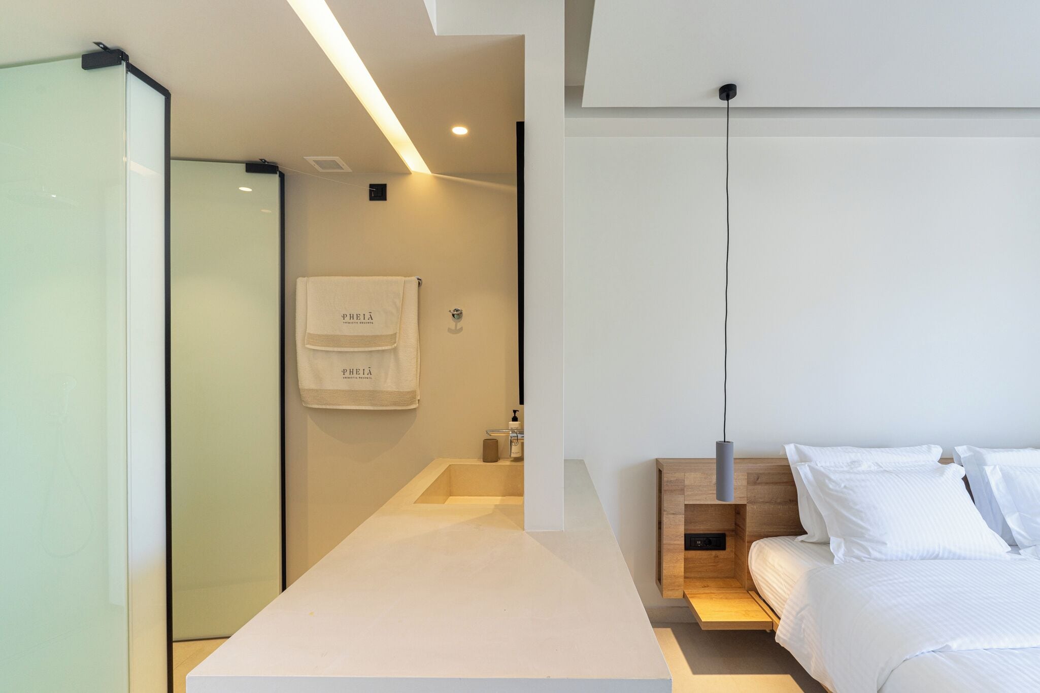 A modern hotel room with a minimalist design, featuring a large bed, a bedside table, and a glass shower enclosure.