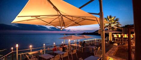 An outdoor dining area with tables and chairs, large umbrellas, and a view of the sea at dusk.