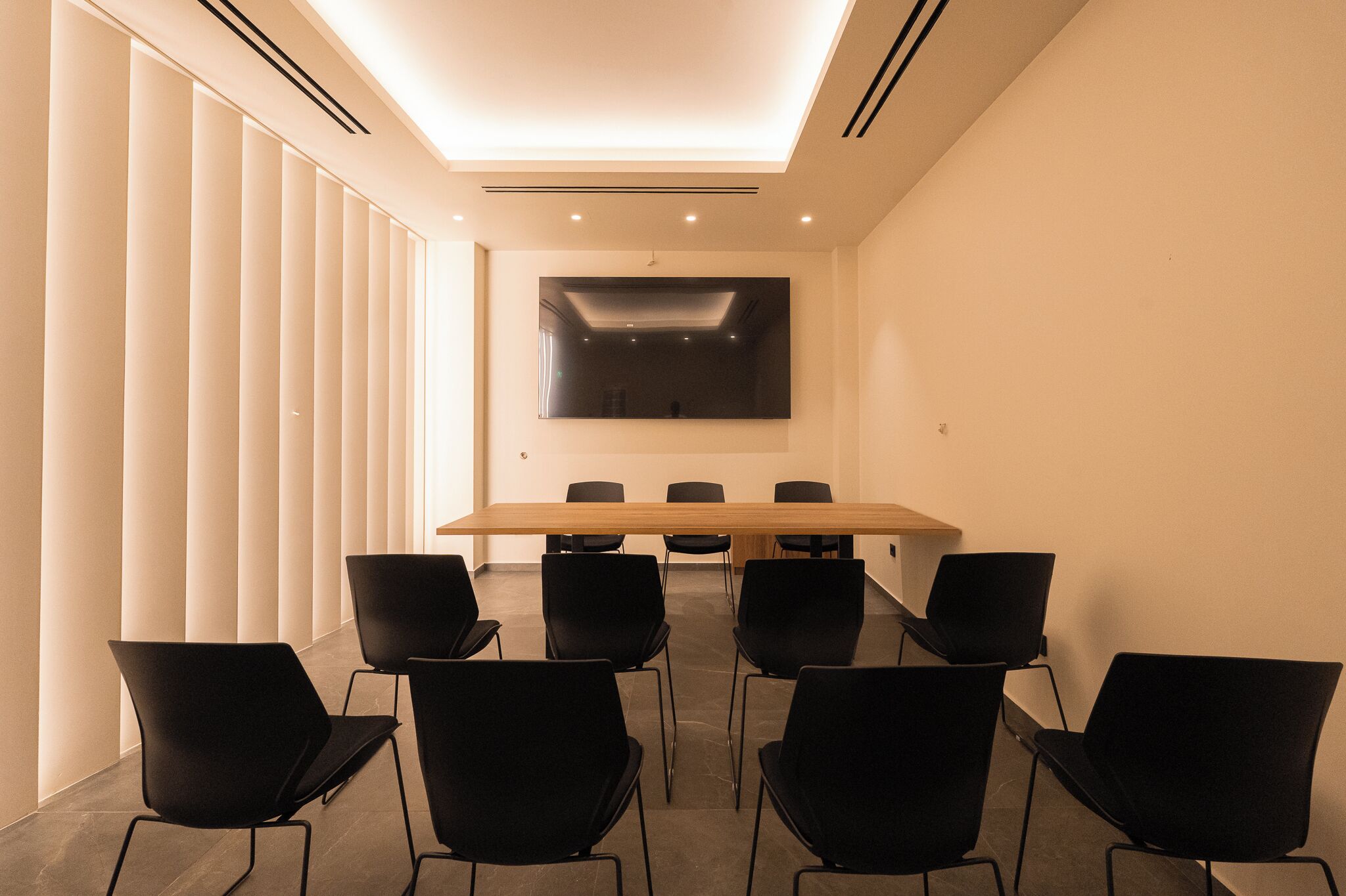 A conference room with a long table and six chairs, a large flat-screen TV on the wall, and vertical blinds on the windows.
