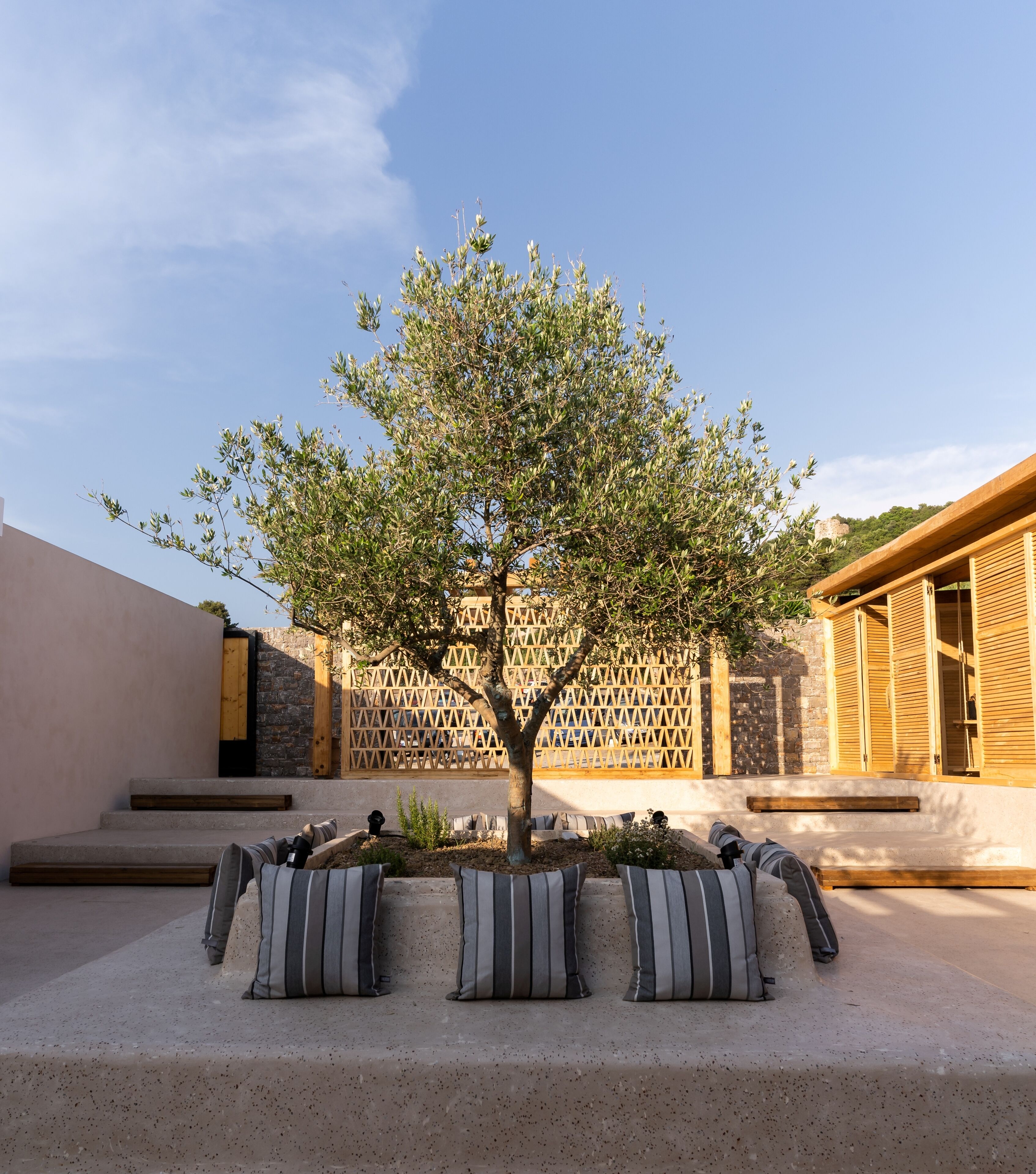A tree in a planter with striped cushions, a stone wall, and a wooden building with shutters.