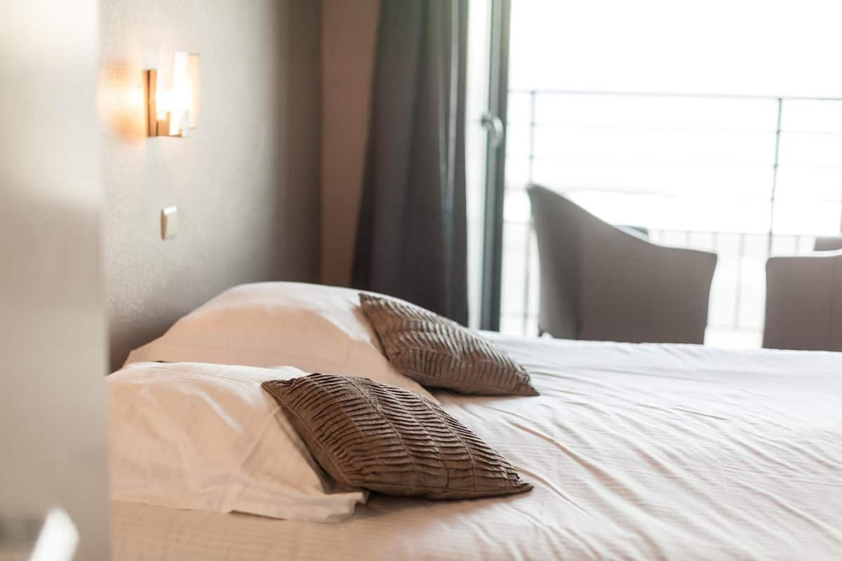 A hotel room with a bed, pillows, a nightstand, and a view of the outside.