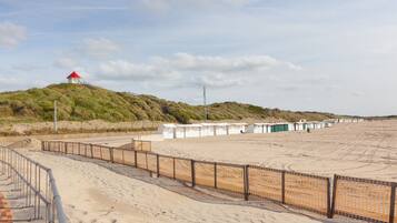 A sandy beach with a fenced walkway, a small hill with a red-roofed structure, and several white buildings in the background.