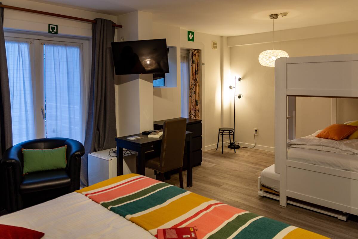 A hotel room with a bed, a desk, a chair, a TV, and a bunk bed.
