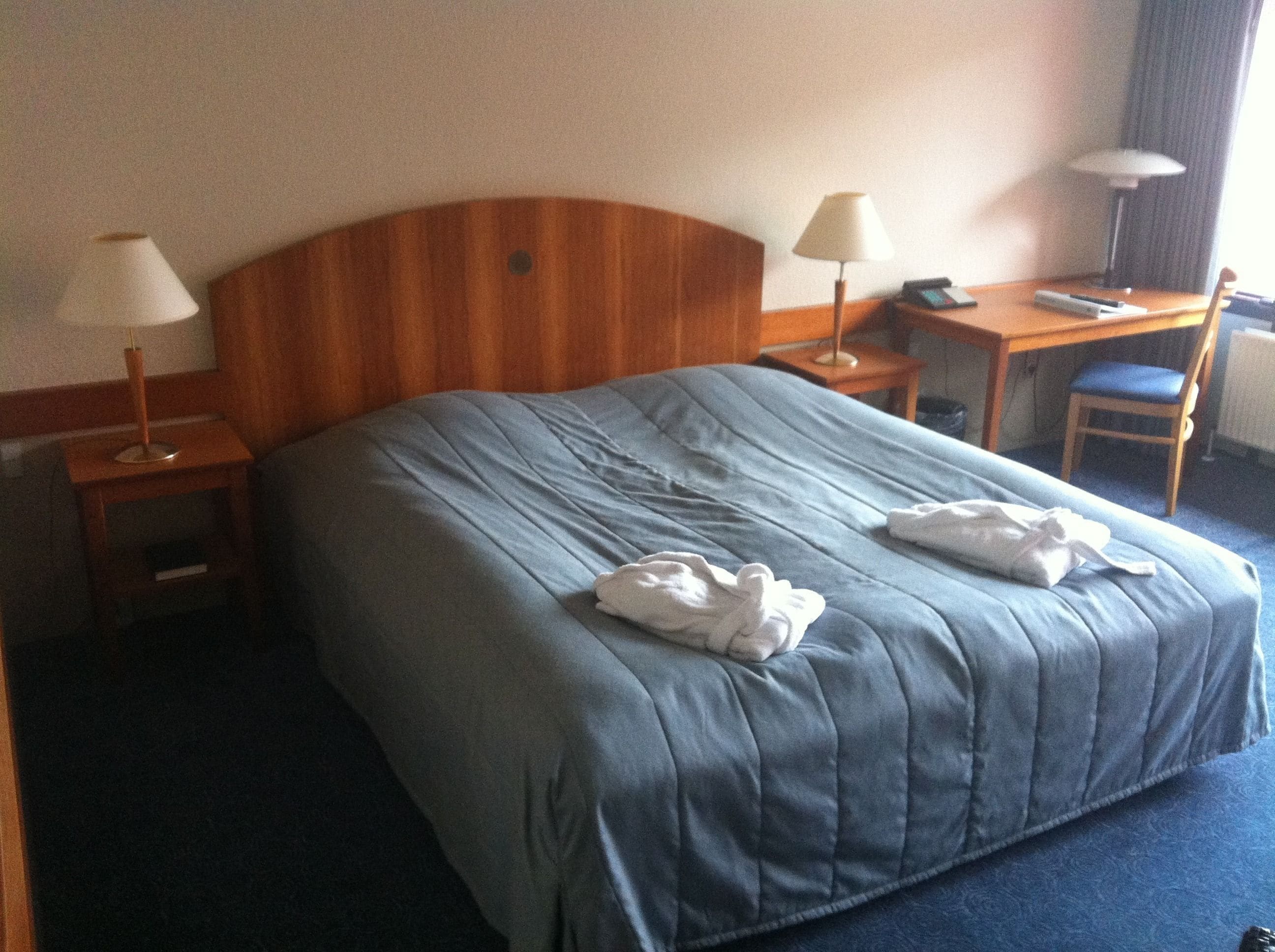 A hotel room with a large bed, two bedside tables with lamps, a desk with a chair, and a window with curtains.