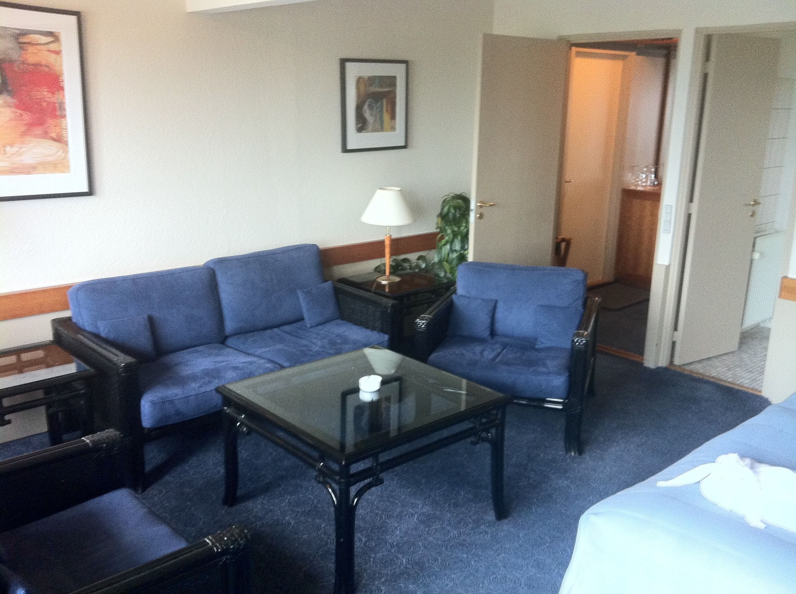 Ein Hotelzimmer mit blauem Sofagarnitur, einem Glastisch, einer Lampe und einer Tür zu einem Badezimmer.