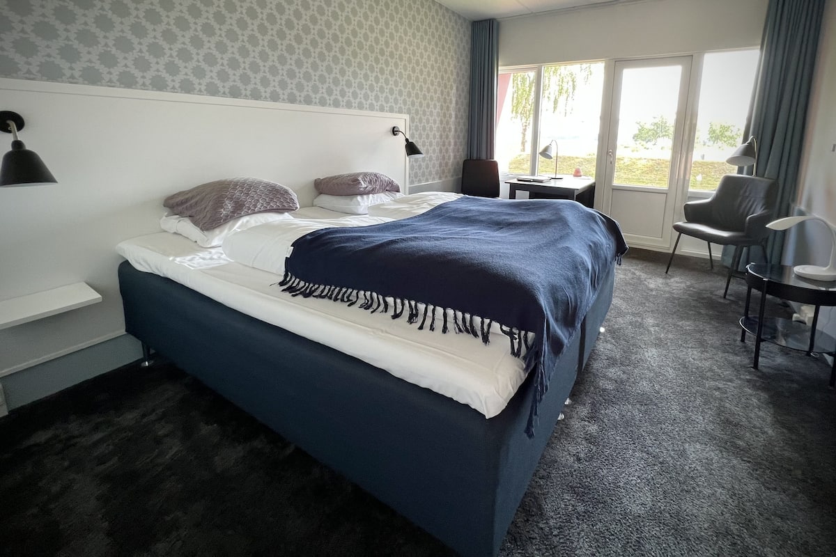Une chambre d’hôtel avec un grand lit, un bureau et une chaise. Il y a une fenêtre avec des rideaux, une lampe fixée au mur et un papier peint à motifs.