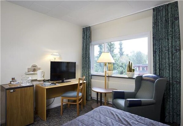 Habitación de hotel con cama, escritorio con televisor, silla, lámpara y vista a árboles por la ventana.