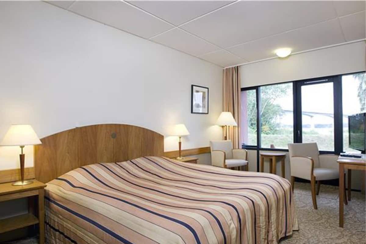 Une chambre d’hôtel avec un grand lit, deux tables de chevet avec des lampes, une chaise, une petite table et une fenêtre donnant sur un beau paysage.