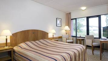 A hotel room with a large bed, two bedside tables with lamps, a chair, a small table, and a window with a view.