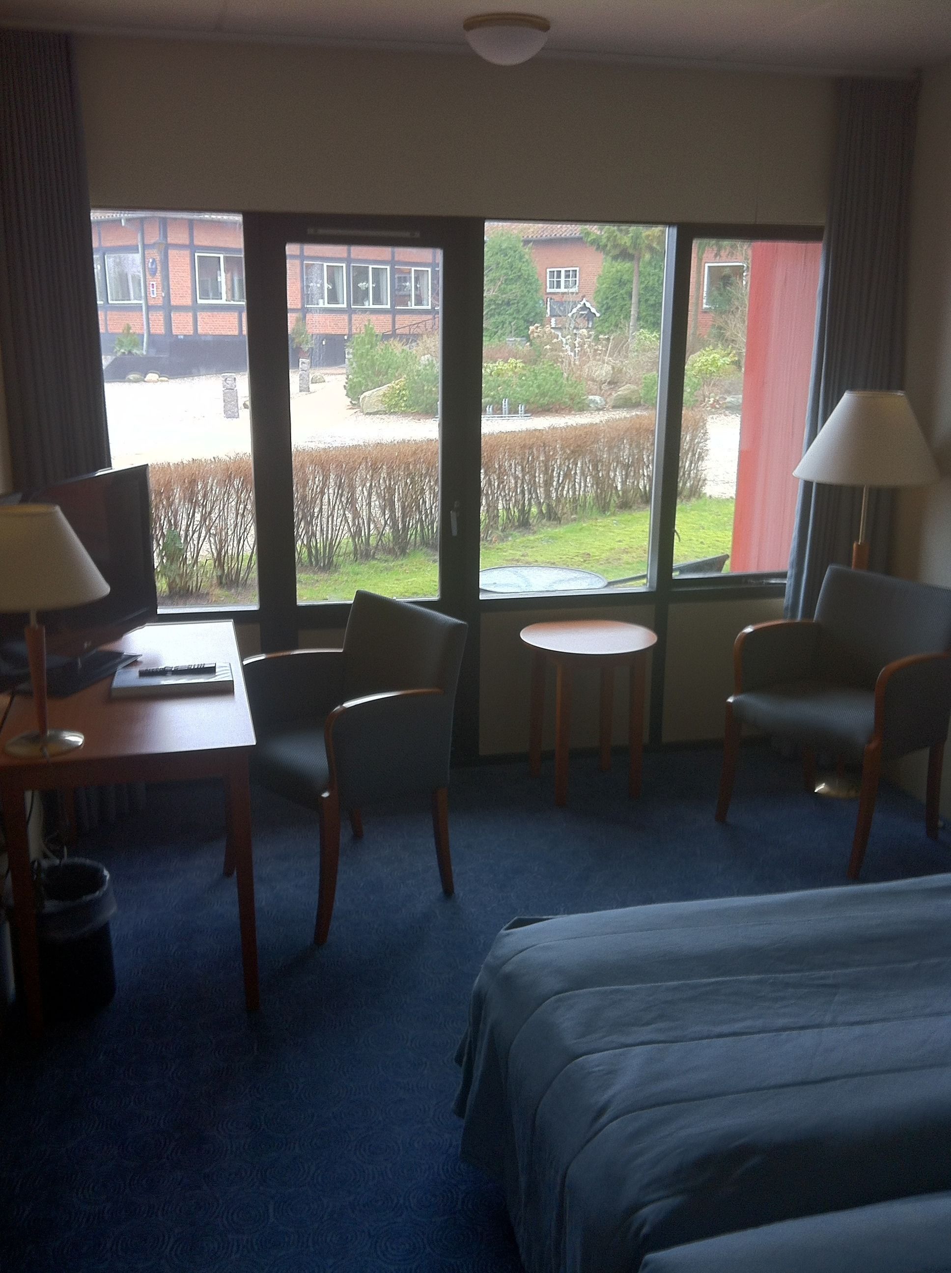 A hotel room with a bed, two chairs, a small table, a desk with a lamp, and a view of a garden and buildings through large windows.