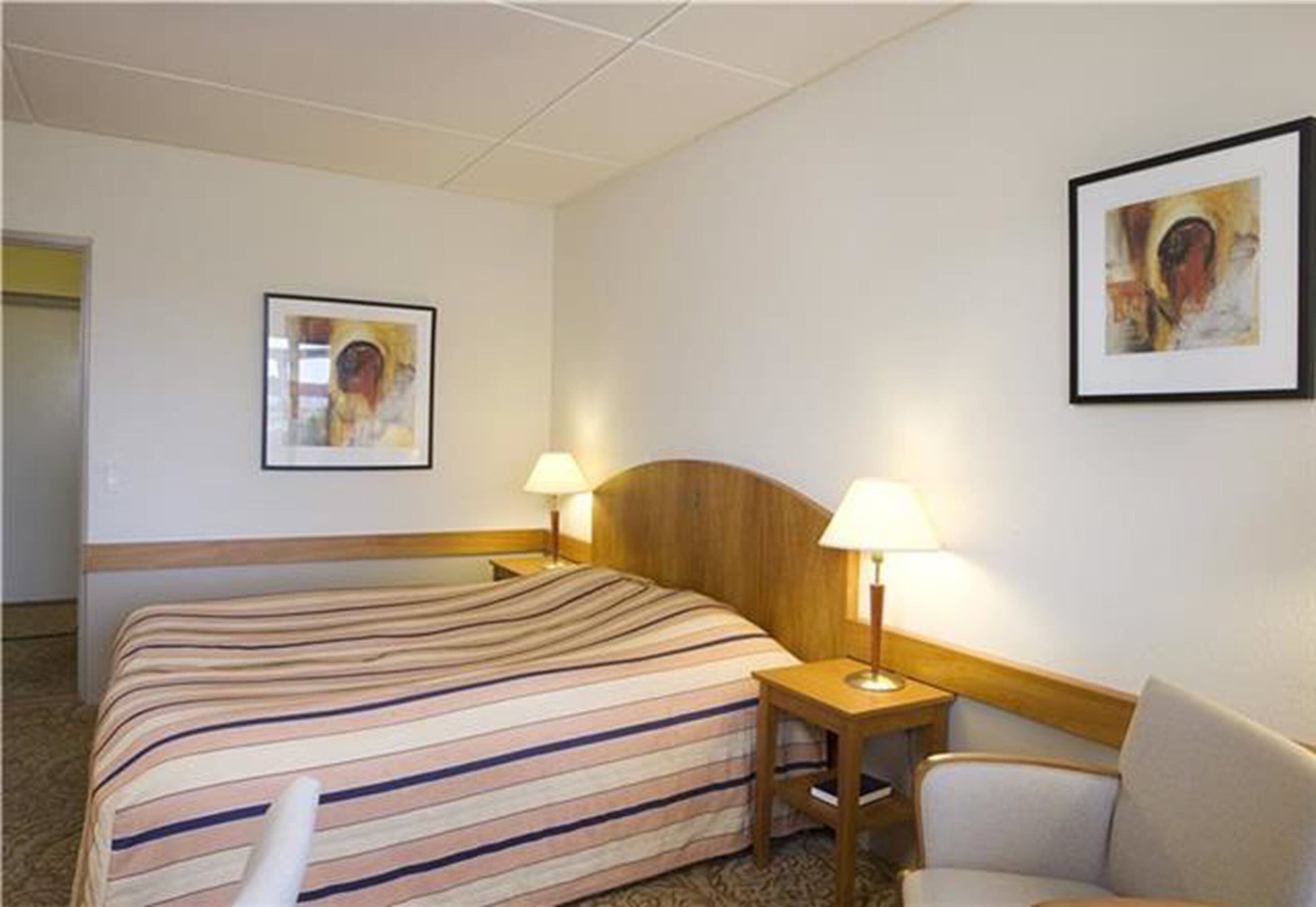 Quarto de hotel com uma cama, duas luminárias de cabeceira, uma cadeira e duas obras de arte emolduradas nas paredes.