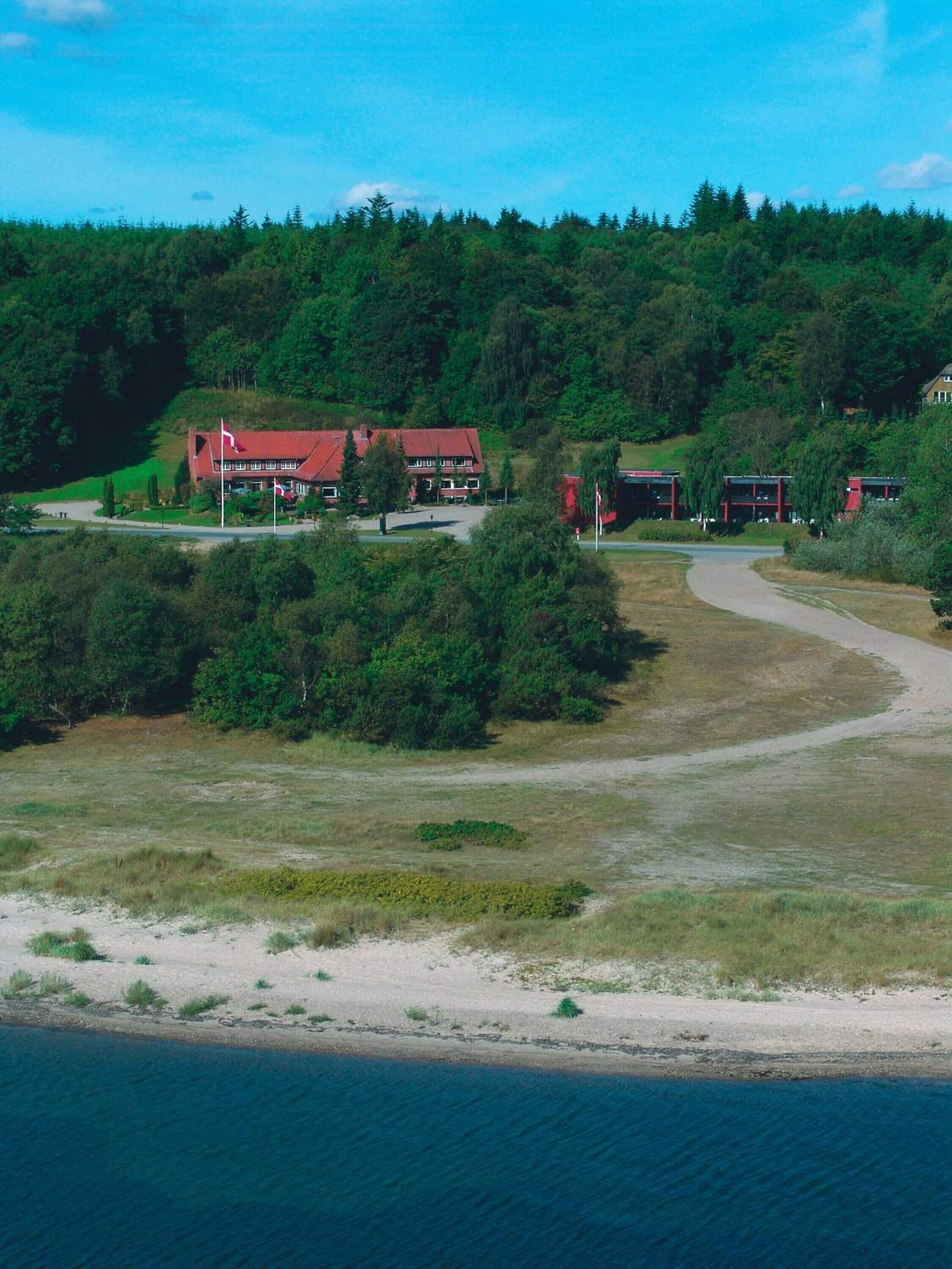Una propiedad costera con un edificio principal y varias construcciones más pequeñas, rodeada de un bosque denso y una playa de arena.