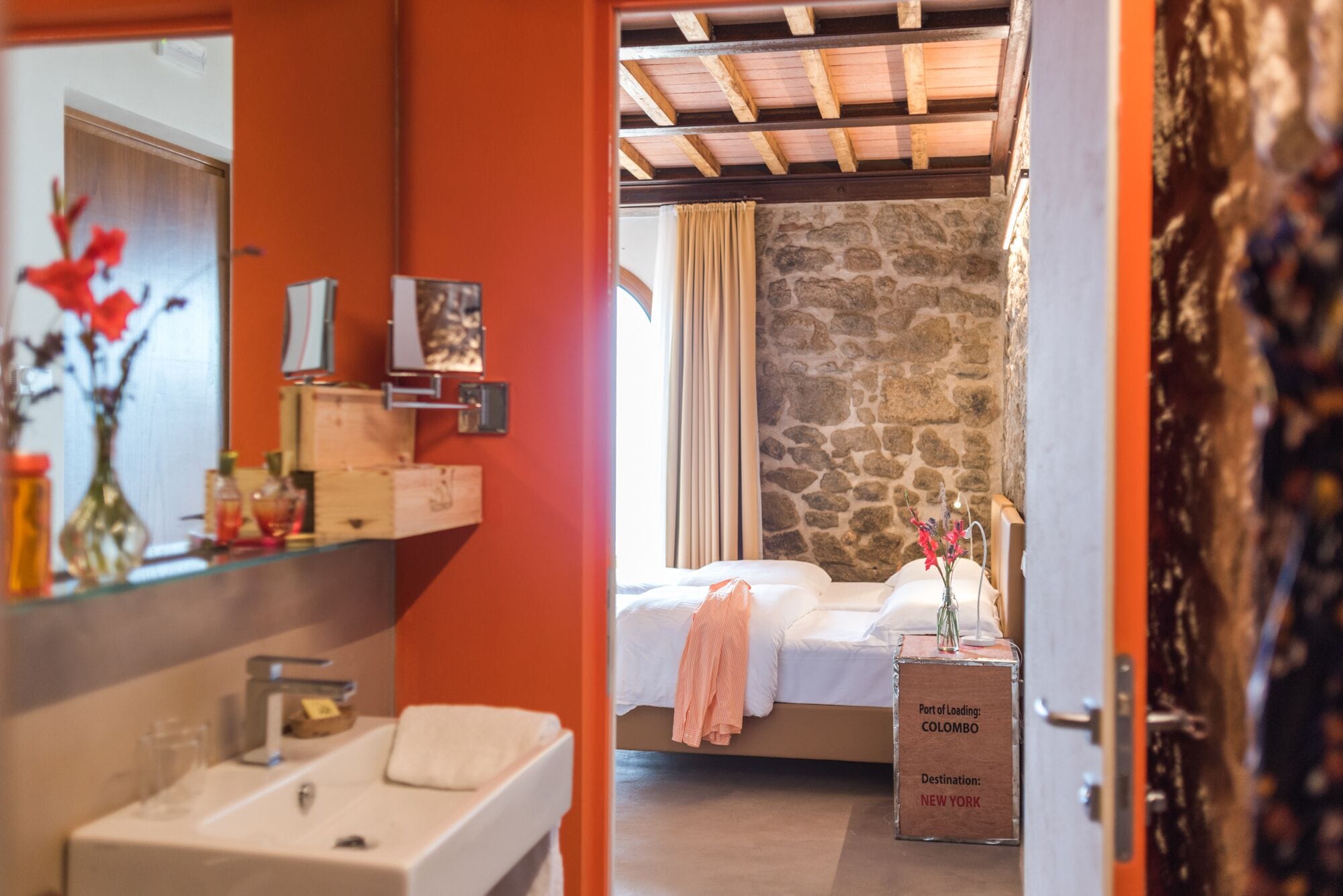 A bedroom with a stone wall, a bed with a pink towel, and a suitcase labeled 'Port of Loading: Colombo, Destination: New York'.