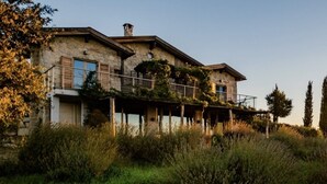 Una casa de piedra rústica con balcón, rodeada de altas hierbas y árboles, al atardecer.