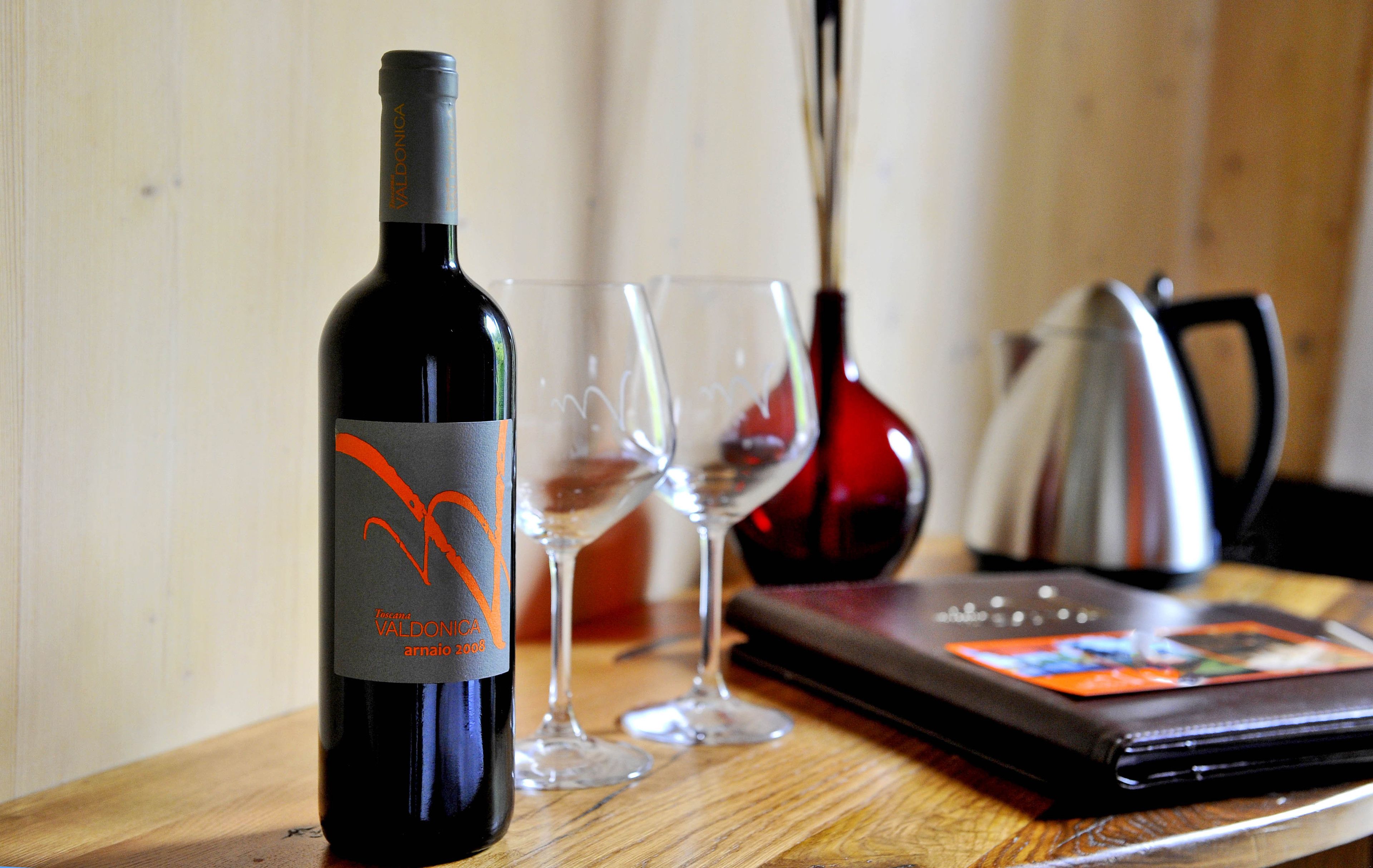 A wine bottle, two wine glasses, a red vase, and a black wallet on a wooden table.