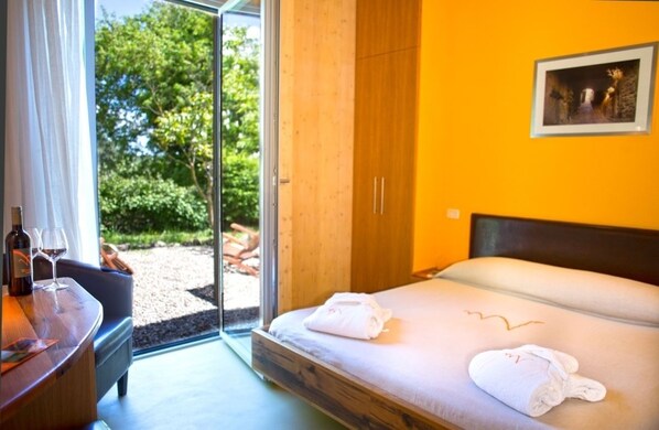 A hotel room with a bed, a desk with a chair, a window with curtains, and a view of greenery outside. - Valdonica Winery & Vineyard Residence (Roccastrada)