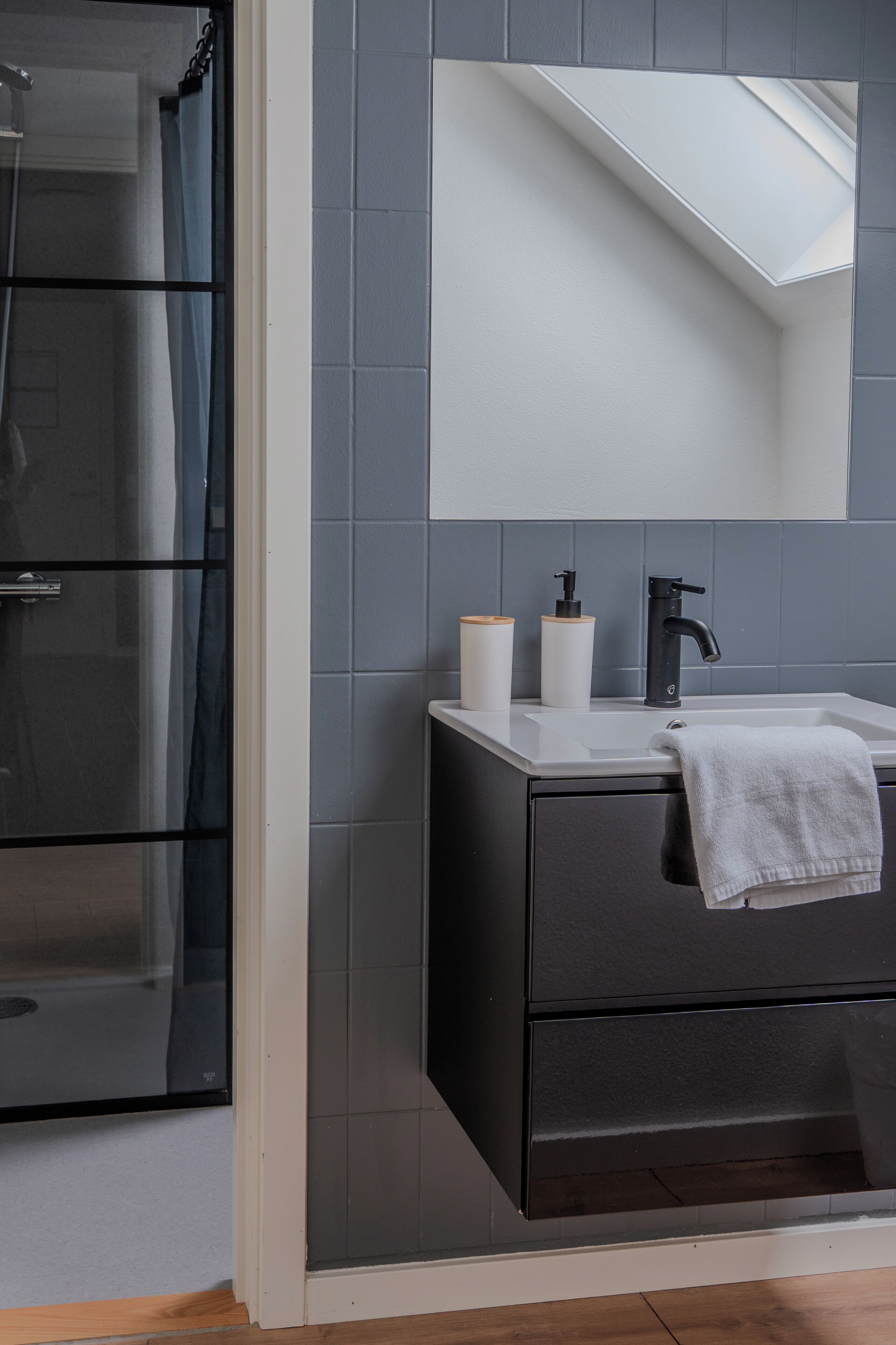 Un baño moderno con un mueble oscuro, un lavamanos blanco, un espejo y una zona de ducha.