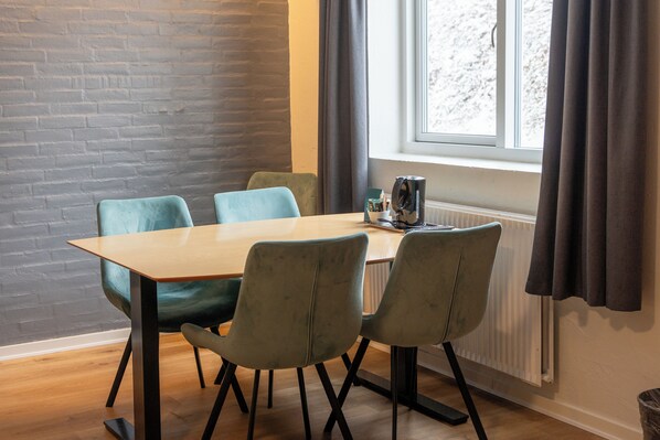 A dining area with a wooden table and four chairs, a radiator, and a window with curtains. - KonceptHotel (Oksbol)