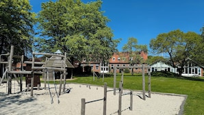 Een speeltuin met klimconstructies en een zandbak, omringd door gras en bomen, met een gebouw op de achtergrond.